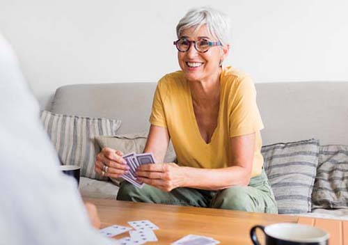 An elderly woman with short gray hair and glasses, wearing a yellow shirt, sits on a couch, smiling while playing cards in a relaxed setting.