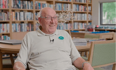 Older person wearing glasses and a beige polo shirt seated in a library with bookshelves and tables in the background