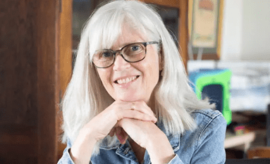 Person with glasses and long grey hair seated indoors, resting hands under chin, with shelves and workspace items in the background