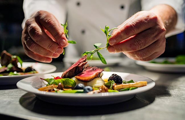 Chef adding garnish to a plated lamb meal on a table