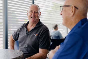 Two older men sit at a table, smiling and conversing. One wears a dark polo, the other a blue shirt. Sunlight filters through window blinds, creating a warm atmosphere.