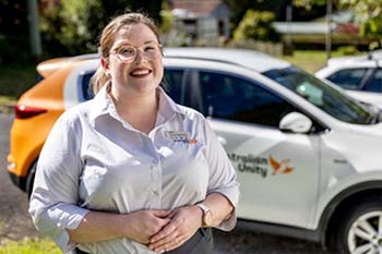 Smiling woman in a white shirt stands outdoors beside a car with an orange and white logo. The scene is sunny and the background is leafy and bright.