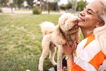 A woman with long blonde hair smiles joyfully as a fluffy dog licks her face in a grassy park. The scene conveys happiness and companionship.