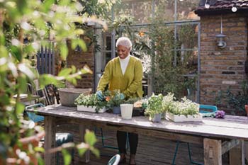 An older woman stands at a wooden table in a sunny garden, tending to potted plants. She smiles, surrounded by greenery and a rustic brick wall.