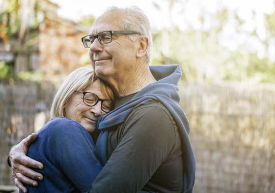 An elderly couple embraces warmly outdoors, both smiling gently. The man wears glasses, and a sweater draped over his shoulders. Cozy, affectionate mood.