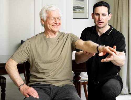 An older man sits and raises his arm, guided by a younger man in a home setting.
