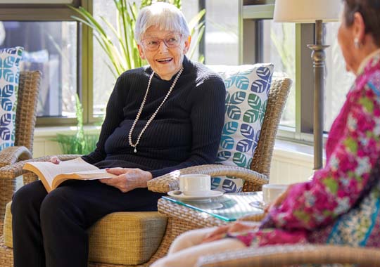 Elderly woman in a black sweater and pearl necklace smiles while holding a book, sitting on a wicker chair with a printed cushion. Another person listens, creating a warm and conversational atmosphere.