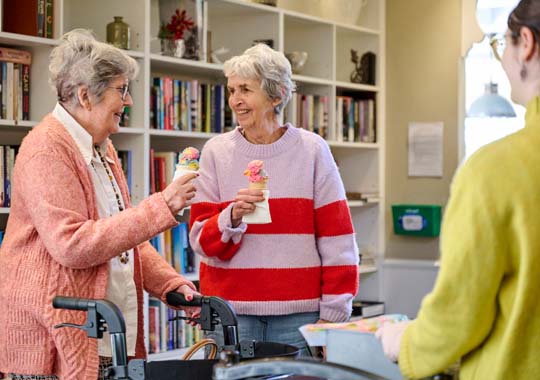 Two elderly women joyfully hold ice cream cones, standing near a bookshelf. A caregiver in the foreground helps, creating a cheerful and supportive atmosphere.