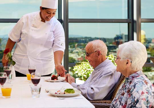 A smiling chef serves dessert to an elderly couple seated at a dining table on a patio.