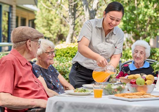 A cheerful caregiver pours juice for three smiling seniors seated at an outdoor table with fruits and snacks. The scene is lively and warm.