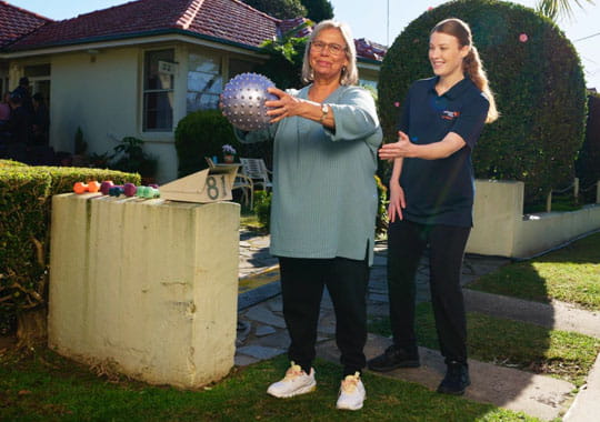 A senior woman and a younger woman outdoors engaging in an exercise routine with a spiky massage ball, smiling, suggesting a sense of support and encouragement.
