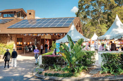 Outdoor community event with white tents and a brick building featuring solar panels. Attendees walk around, engaging in festive activities amidst greenery.