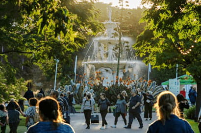 A bustling park scene at sunset with people walking and gathering near a large, ornate fountain surrounded by lush greenery.