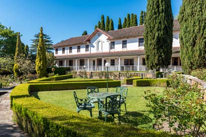 A charming two-story building with white walls and red-tiled roof is surrounded by manicured gardens. Green metal table and chairs sit on a lawn under clear blue skies.