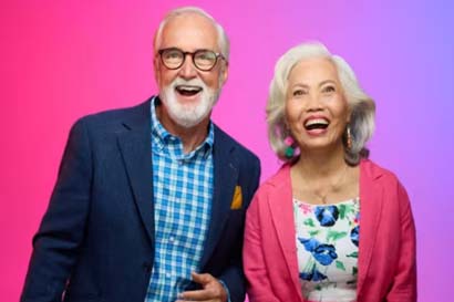Elderly man and woman smiling against a vibrant pink and purple background. He wears glasses and a blazer; she wears a floral dress and pink cardigan. 