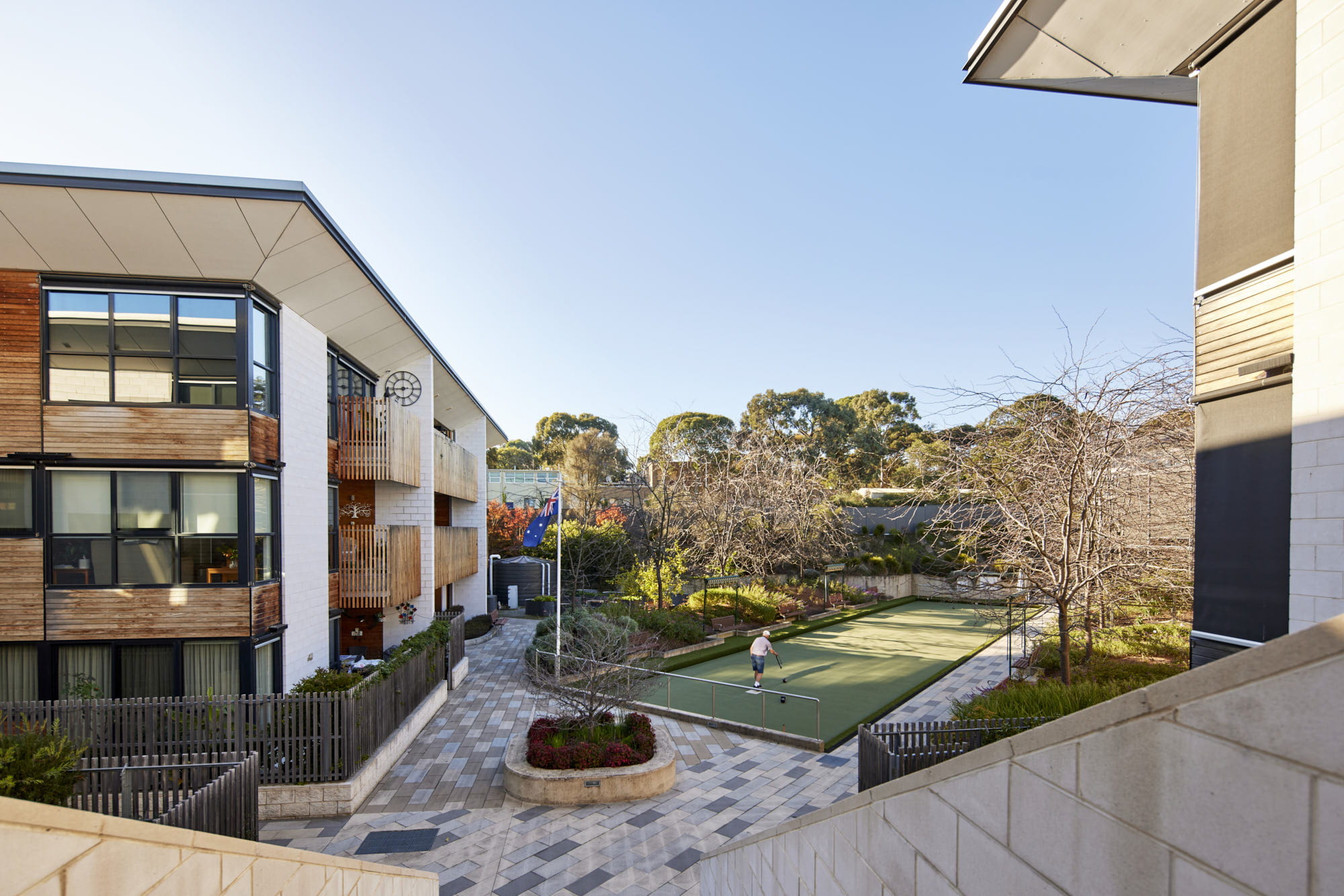 A person plays on a small outdoor sports court between modern buildings and landscaped gardens on a sunny day.