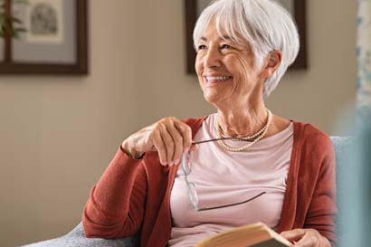 Elderly woman with short white hair smiling while holding glasses and a book, wearing a red cardigan and pearls.