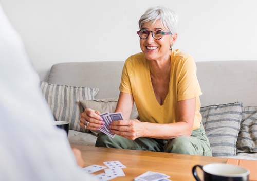 Elderly woman with short gray hair and glasses, smiling while playing cards at a table. Relaxed atmosphere with a sofa and striped pillows in the background.