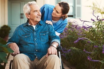 A retiree in a wheel chair looks at flowers while their carer smiles.
