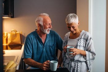 Retired couple in a warmly lit kitchen, smiling while they drink tea.