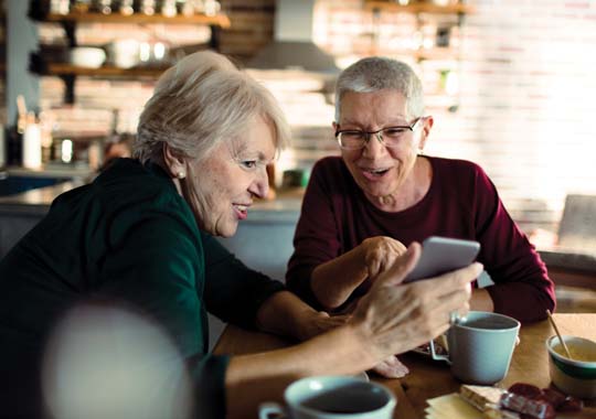 Two older women sitting at a table, focused on a cell phone, sharing a moment of connection and conversation.