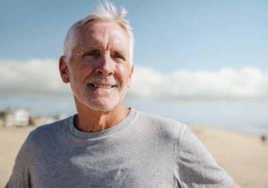 Smiling older man with short gray hair in a gray shirt stands on a sunny beach. Blurred buildings and cloudy sky are in the background.