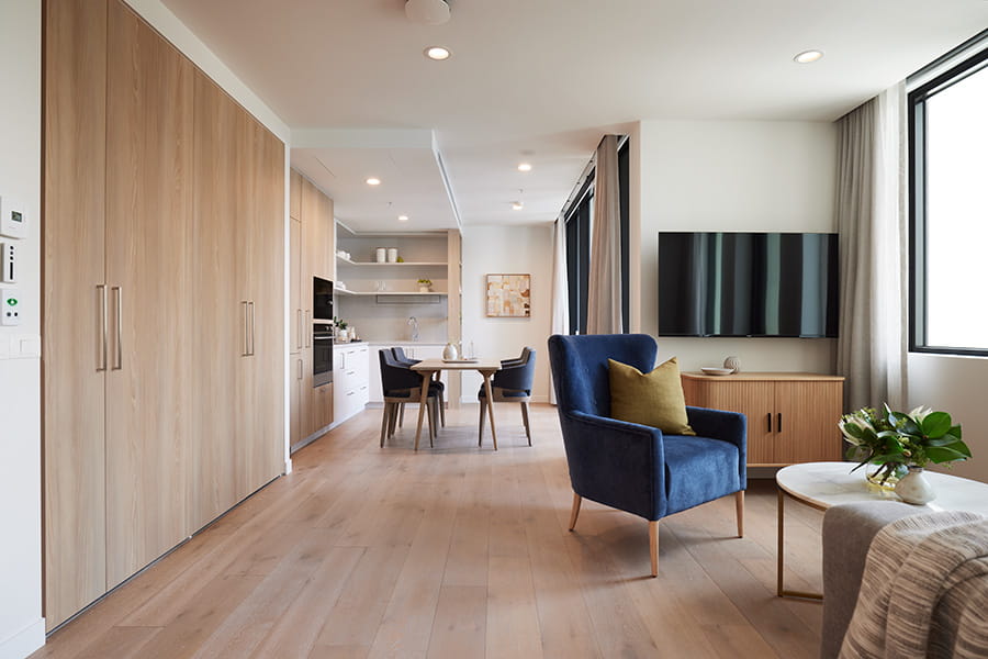 Stylish apartment interior with light wood flooring, a blue armchair, wall-mounted TV, and an open kitchen and dining area in the background. 	