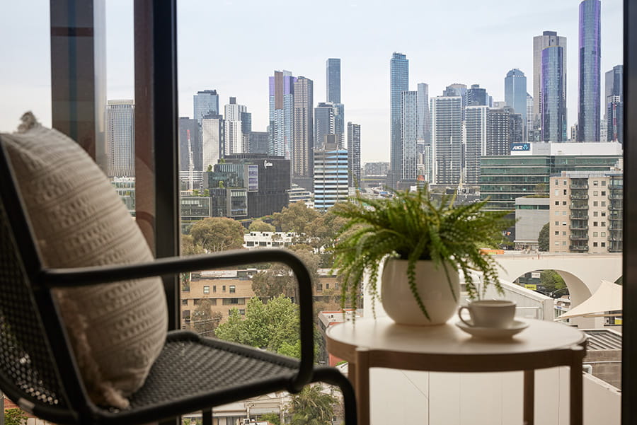 View of a modern city skyline through a large window, with a chair, cushion, potted fern, and coffee cup on a small round table in the foreground. 	