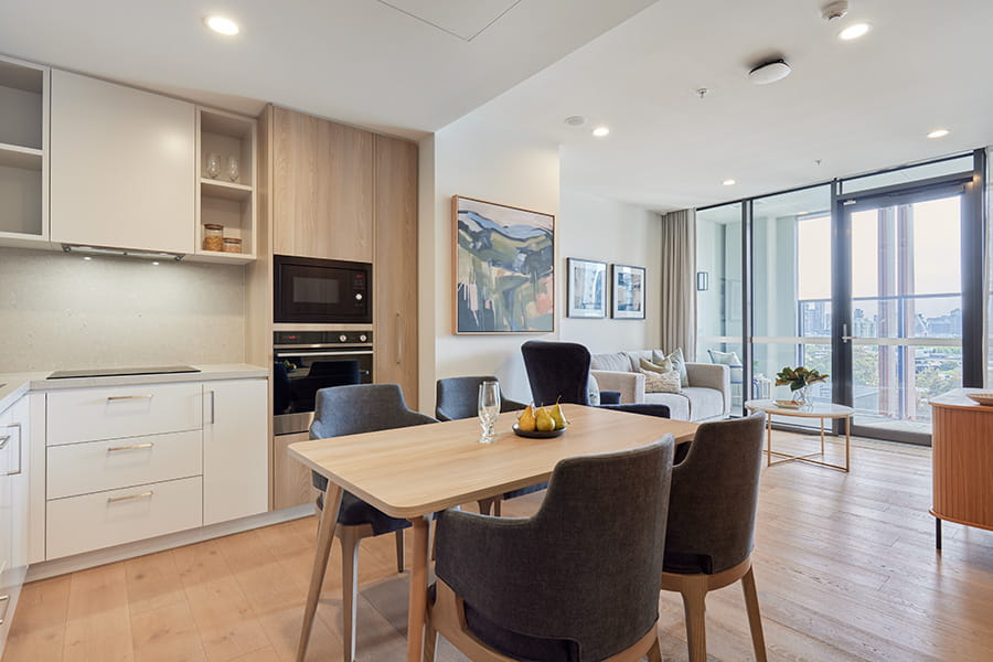 Contemporary open-plan apartment featuring a dining table with four chairs, a modern kitchen with light wood cabinetry, and a living area with sofa and glass doors opening to a balcony overlooking the city.