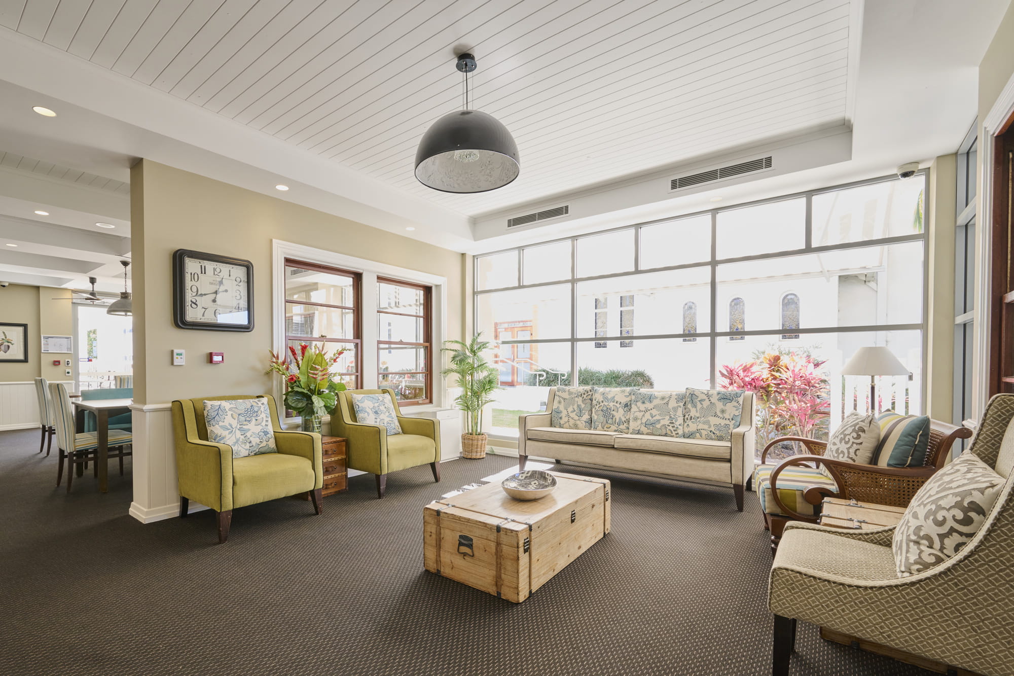 Bright living room with natural light from large windows. Features two green armchairs, a floral sofa, wicker chairs, and a wooden chest as a coffee table. 