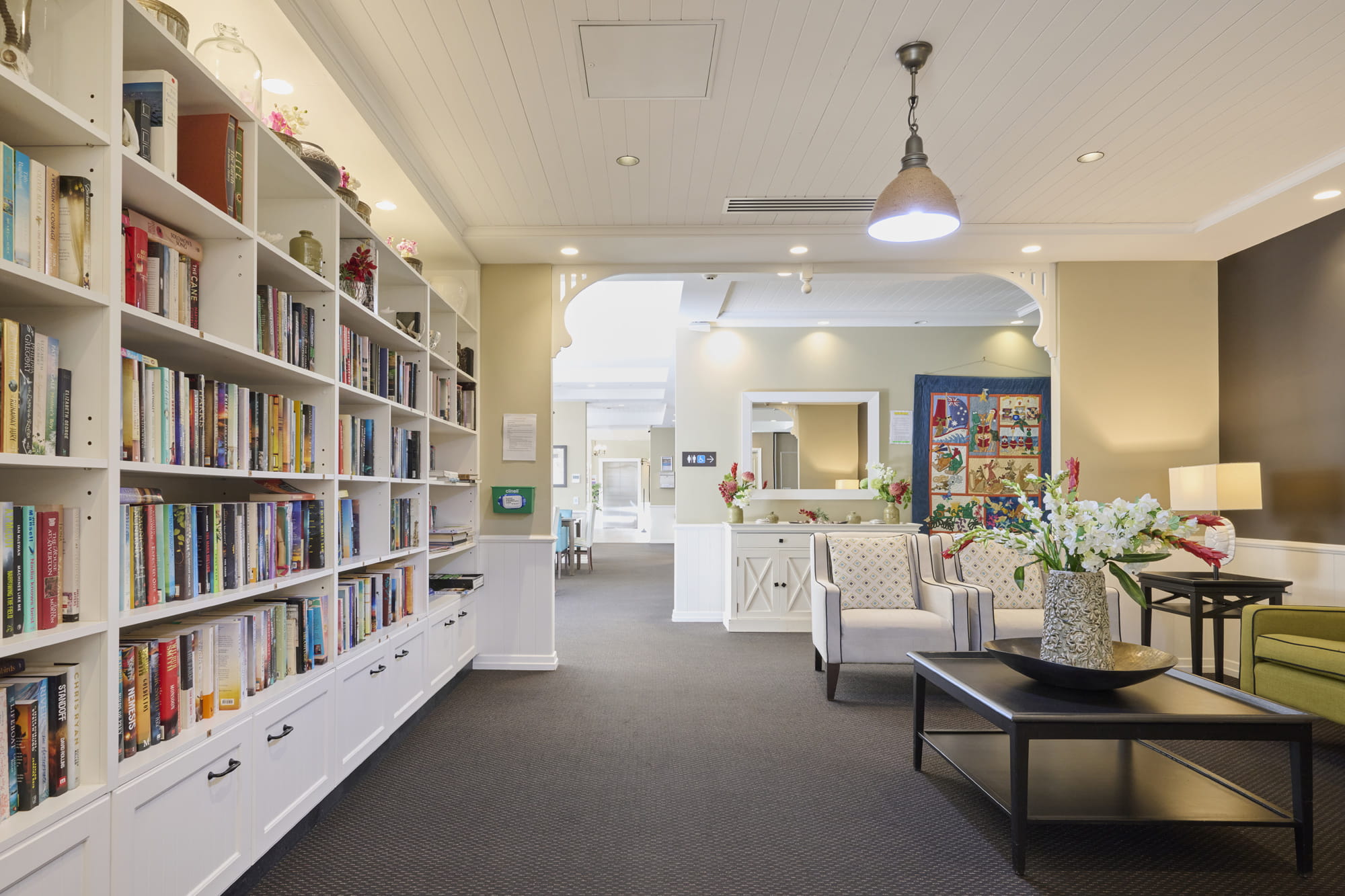 Spacious room with white bookshelves on the left, colorful books. Modern decor includes cozy armchairs, a central table with flowers, and soft lighting. 