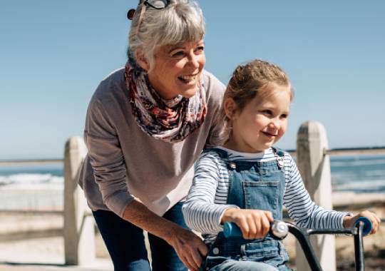 A grandmother pushing her granddaughter on a bike next to the beach.