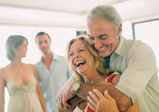 Older couple laughing and embracing in a sunny room with their adult children in the background.