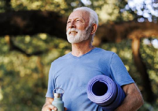 Older man in a park, with a rolled-up yoga mat under one arm and a water bottle in his other hand.