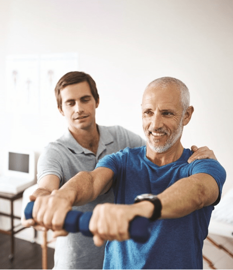A gray-haired man in a blue shirt smiles while doing arm exercises with dumbbells. A younger man in a gray shirt assists him, creating a supportive atmosphere.
