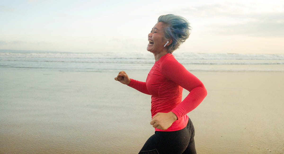 An older woman running on the beach grinning as the wind combs through her hair.