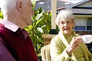 An elderly couple sits outdoors on a sunny day, engaging in a pleasant conversation. The woman, holding a teacup, smiles warmly at the man.