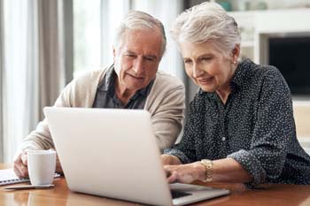 Elderly couple smiling, seated at a table, using a laptop together in a bright room. They appear engaged and content, with a coffee mug nearby.