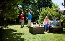 Two women sitting on an outdoor lounge in backyard, two mean standing and talking in the distance