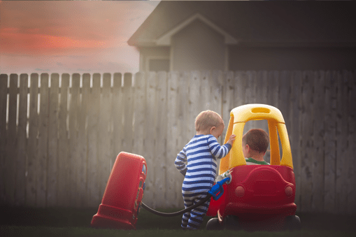 Twin toddlers play with plastic toy cars in their backyard on a late spring/early summer evening.