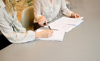 Two mean sitting at a desk looking over paperwork. One woman is writing