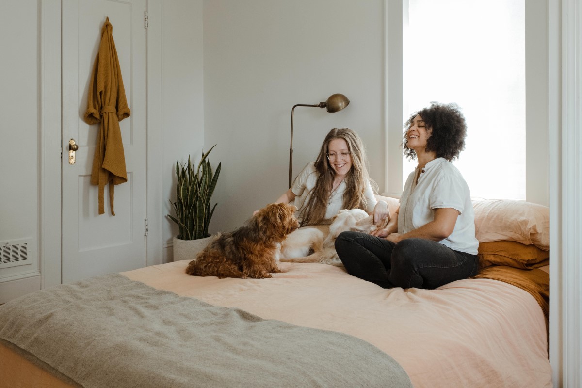 Two women sitting on a bed, laughing with their dog