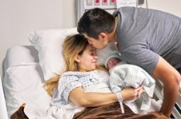 Woman and newborn in hospital bed, her partner kisses her forehead