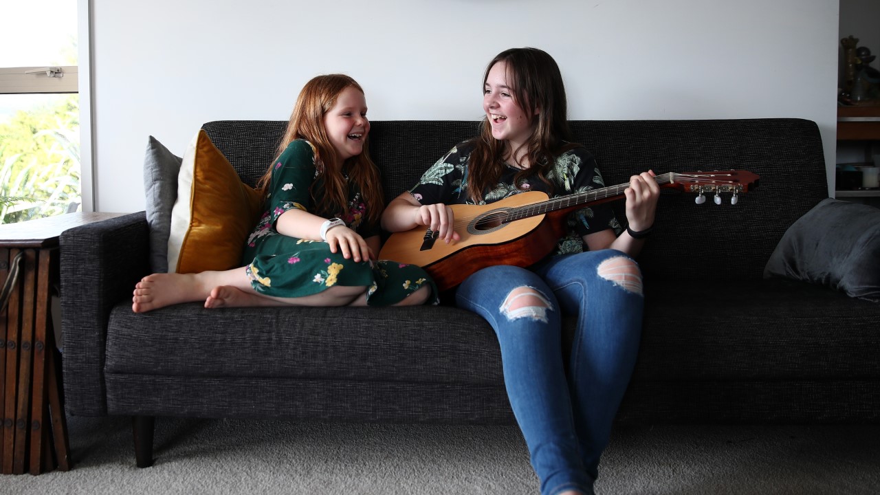 Happy girl with guitar on couch, younger girl next to her smiling
