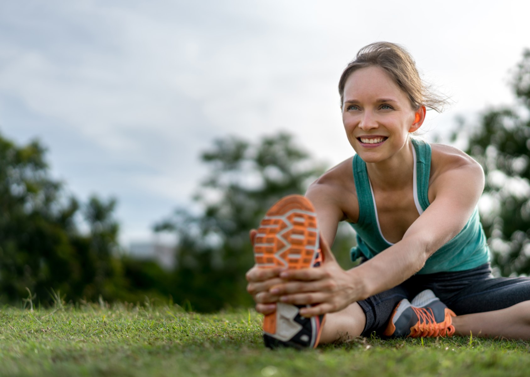 Woman outdoors, sitting on grass and stretching her right leg.