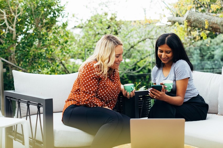 Two women, sitting on outdoors couch, smiling and looking at phone screen