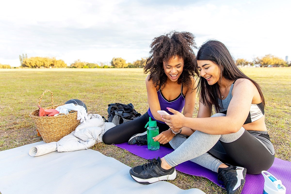 Two women, sitting on exercise mat in park, smiling and looking at phone screen