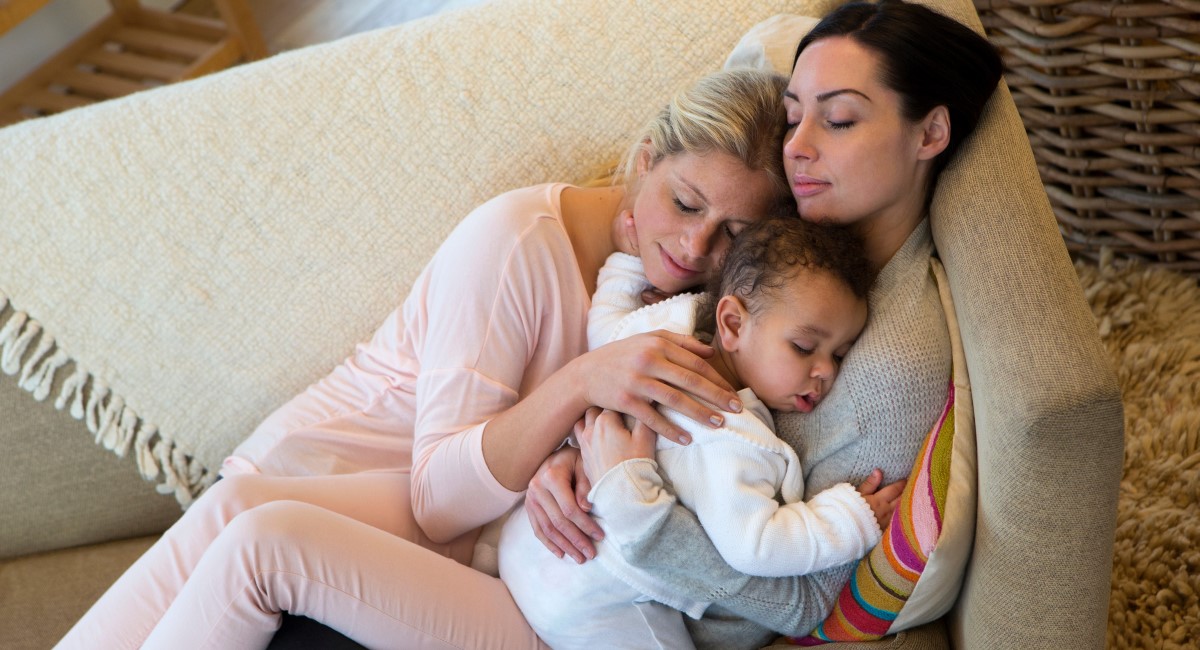 Two women lie sleeping on the couch with their baby