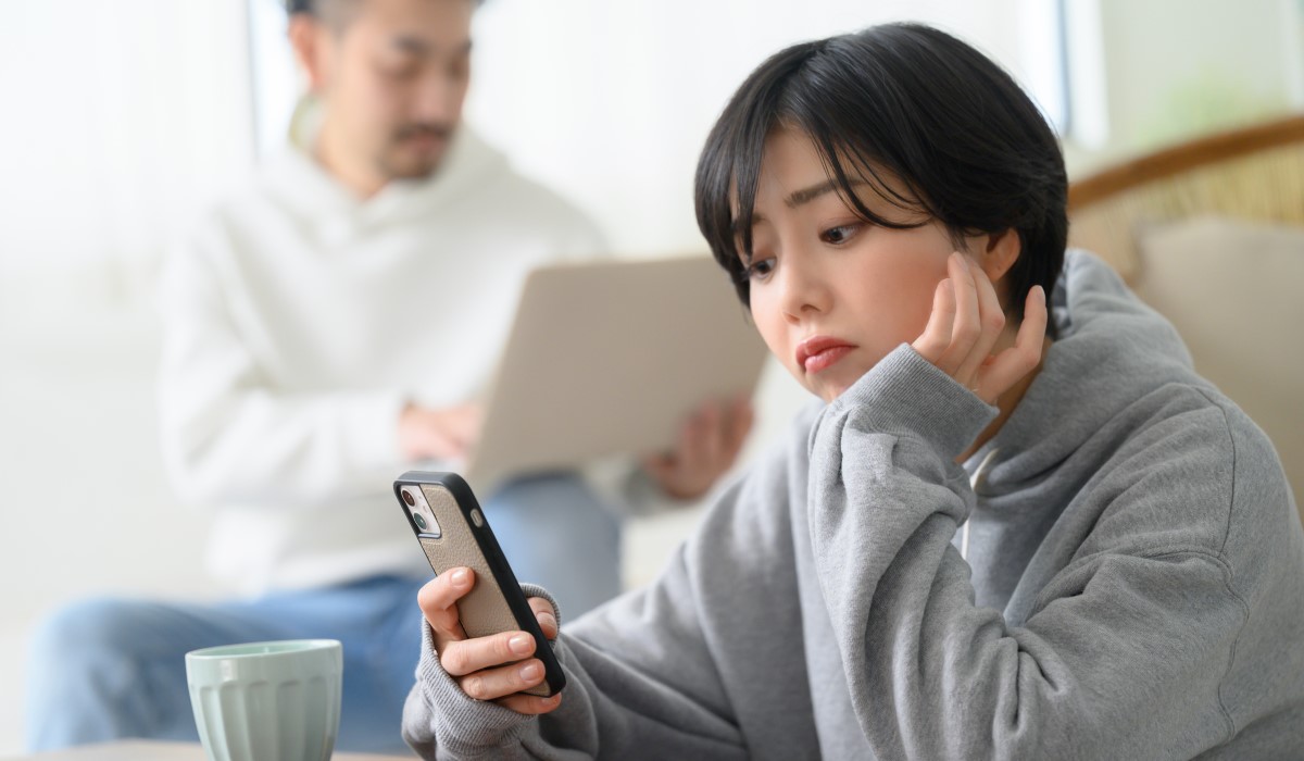 Woman looking lonely and stressed, looking at her phone with male partner looking at laptop in background
