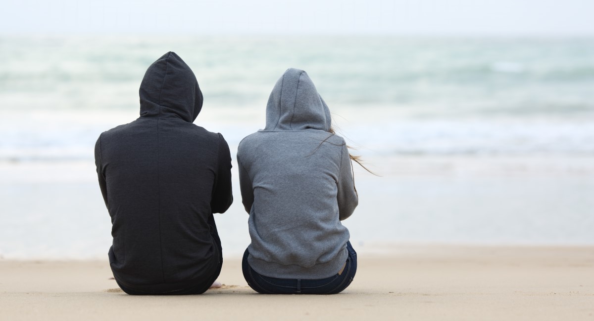 Man and woman in hoodies sitting on beach with backs to camera
